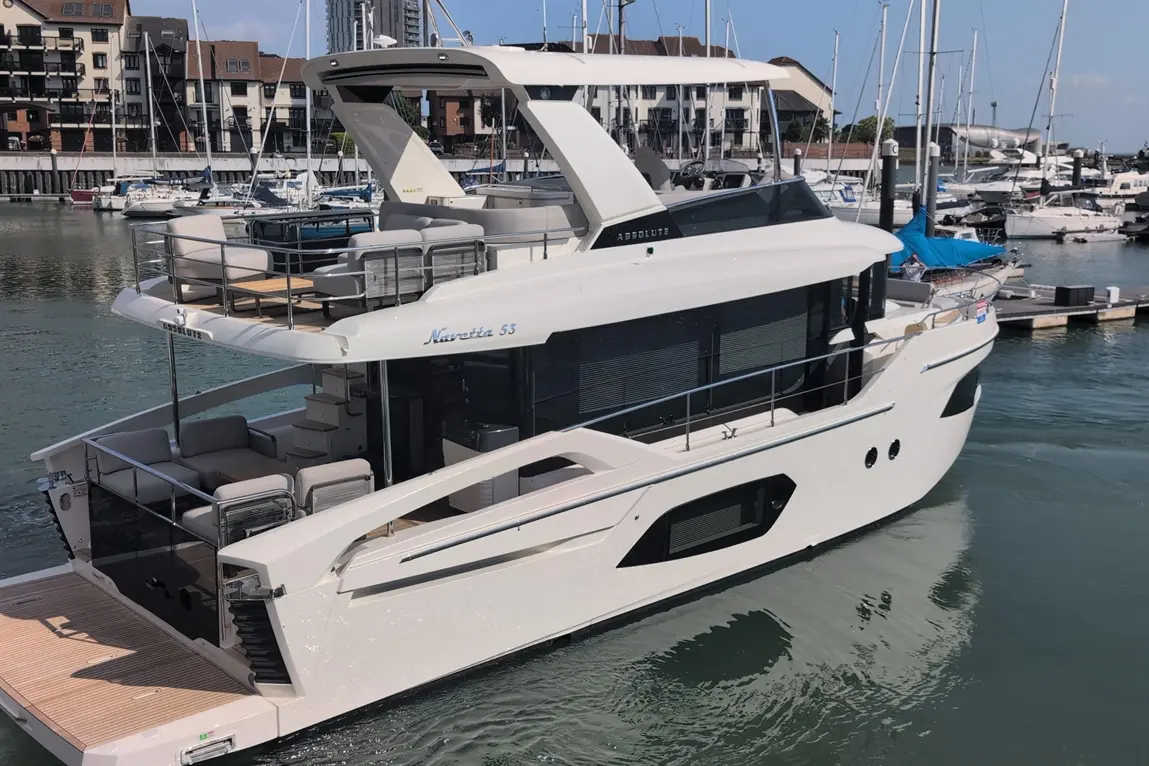 A sleek white motor yacht, the Absolute Navetta 53, docked at a marina surrounded by smaller boats and buildings.
